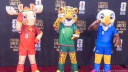 Three 2026 World Cup mascots pose in front of a FIFA 'Football Unites the World' backdrop.