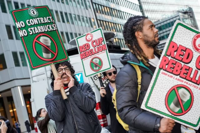 Starbucks employees chant while picketing outside a New York City store in 2025, holding signs reading No Contract No Starbucks and Red Cup Rebellion