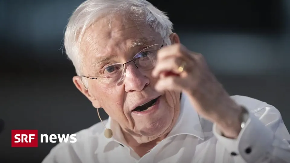 Christoph Blocher speaks into a microphone while gesturing during a public appearance.