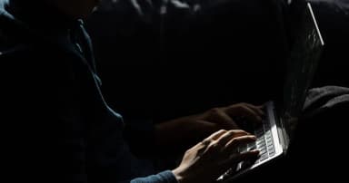 Hände auf einer Laptop-Tastatur in einem dunklen Raum neben einem teilweise geöffneten Bildschirm.