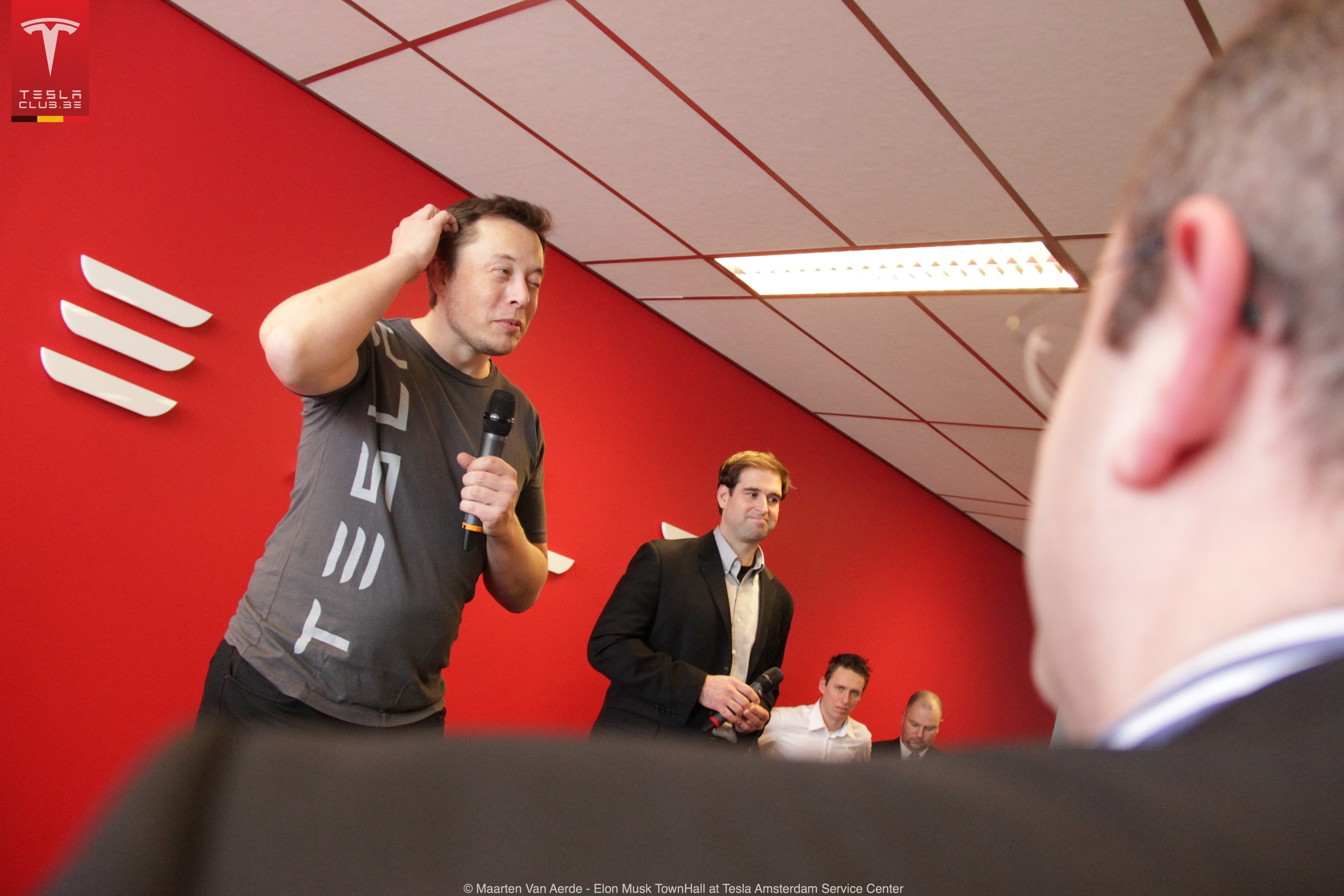 Elon Musk speaking at a public event, gesturing with his hands while addressing an audience