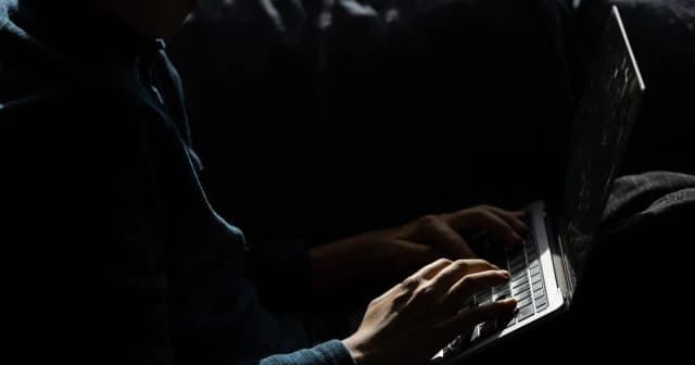 Hands on a laptop keyboard in a dark room beside a partially open computer screen.